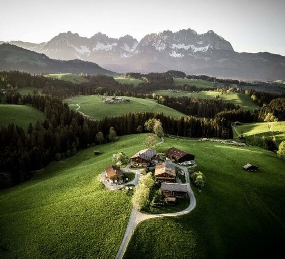 Luftaufnahme eines Bauernhofs auf einer grünen Wiese vor einem bewaldeten Tal und schroffen Berggipfeln.