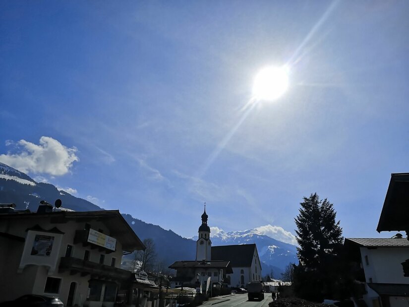 Blick auf eine Dorfkirche mit Zwiebelturm vor schneebedeckten Bergen unter strahlender Sonne.