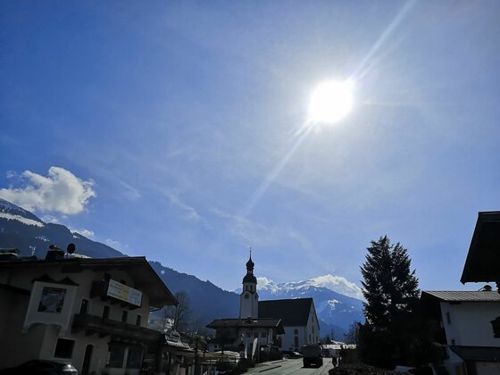 Blick auf eine Dorfkirche mit Zwiebelturm vor schneebedeckten Bergen unter strahlender Sonne.