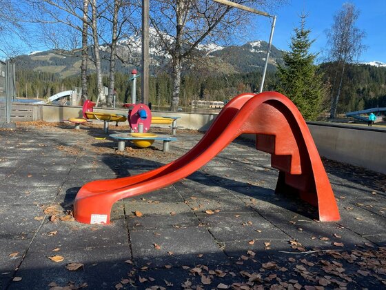 Rote Kinderrutsche auf einem Spielplatz mit Gummiboden vor bewaldeten Bergen.