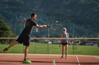 Zwei Personen beim Tennisspielen auf einem Außenplatz vor bewaldeten Bergen.