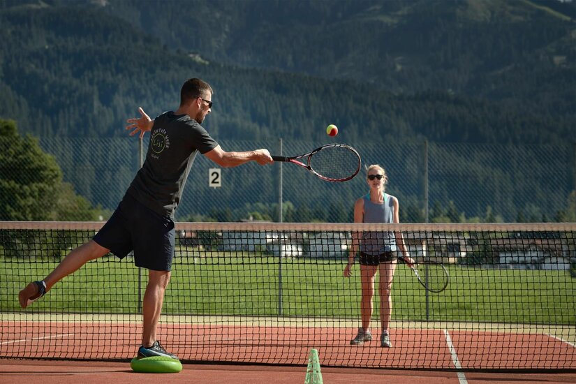 Zwei Personen beim Tennisspielen auf einem Außenplatz vor bewaldeten Bergen.