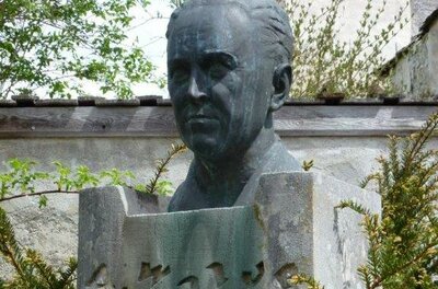Alfons Walde Büste Eine Büste eines Mannes steht auf einem Steinsockel. Um die Skulptur herum sind grüne Pflanzen und Bäume zu sehen.