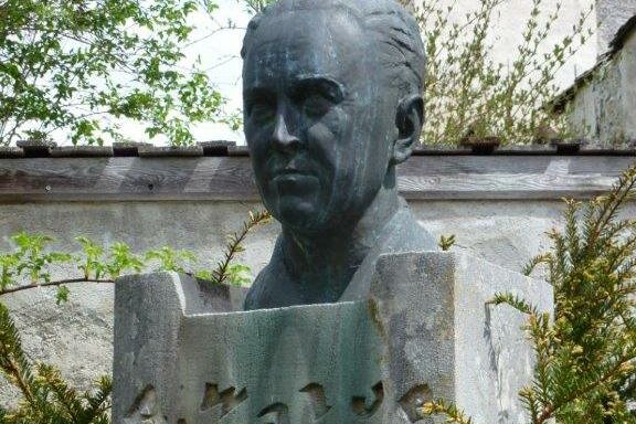 Alfons Walde Büste Eine Büste eines Mannes steht auf einem Steinsockel. Um die Skulptur herum sind grüne Pflanzen und Bäume zu sehen.