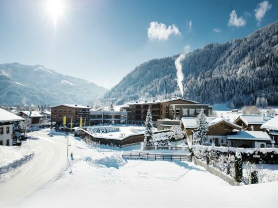 Verschneite Hotelanlage Kitzbühel Lodge vor bewaldeten Bergen bei Sonnenschein.