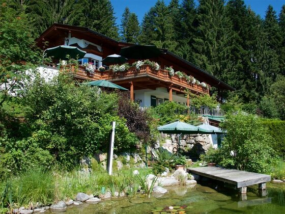 Alpines Holzhaus mit blumengeschmücktem Balkon, Sonnenschirmen und einem kleinen Badeteich im Vordergrund.
