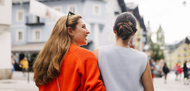 Zwei Frauen spazieren fröhlich durch die malerische Innenstadt von Kitzbühel, umgeben von bunten Gebäuden und Menschen.