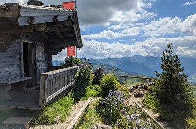 Rustikale Holzhütte mit Terrasse vor einem Bergpanorama unter bewölktem Himmel.
