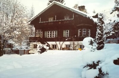 Villa Momo im Winter Zweistöckiges Holzhaus mit Balkon und weißem Sockel in einer verschneiten Gartenlandschaft.