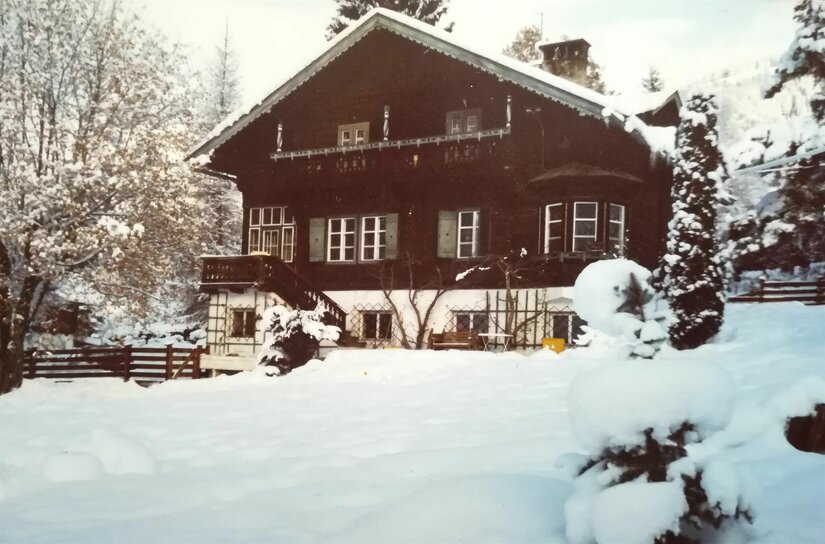 Zweistöckiges Holzhaus mit Balkon und weißem Sockel in einer verschneiten Gartenlandschaft.