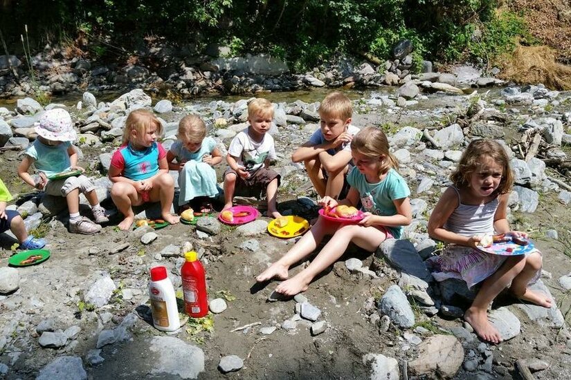 Kinderkunterbunt Eine Gruppe Kinder sitzt am Ufer eines kleinen Baches und genießt ihr Essen. Sie haben bunte Teller und scheinen eine schöne Zeit in der Natur zu verbringen.