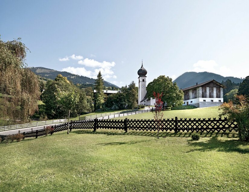 Kirche mit Zwiebelturm und Bauernhaus hinter einem Holzzaun auf einer grünen Wiese vor Bergkulisse.