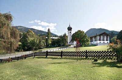Kirche mit Zwiebelturm und Bauernhaus hinter einem Holzzaun auf einer grünen Wiese vor Bergkulisse.