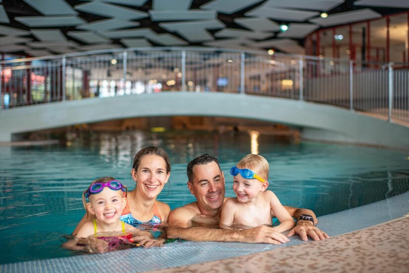 Familie im Badezentrum Aquarena Eine vierköpfige Familie mit zwei Kindern im Wasser eines Hallenbades vor einer Brücke.