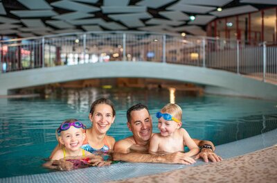 Familie im Badezentrum Aquarena Eine vierköpfige Familie mit zwei Kindern im Wasser eines Hallenbades vor einer Brücke.
