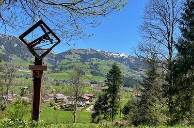 Station am Kreuzweg Ein hölzernes Kreuzweg-Relief im Vordergrund vor einer alpinen Tal- und Berglandschaft bei blauem Himmel.