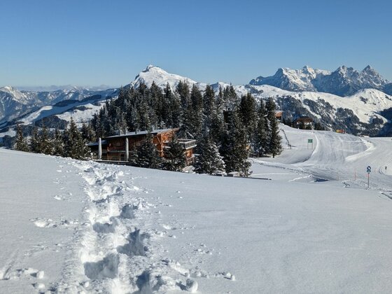 Schneeschuhspur im tiefen Schnee führt zu einer Holzhütte vor verschneitem Bergpanorama.