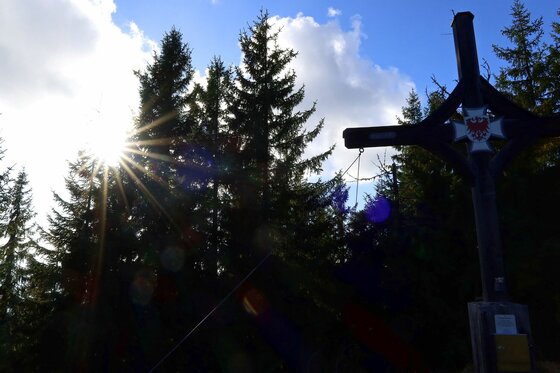 Gipfelkreuz am Kaiserblickweg Gipfelkreuz im Gegenlicht vor Nadelbäumen und blauem Himmel mit Wolken.