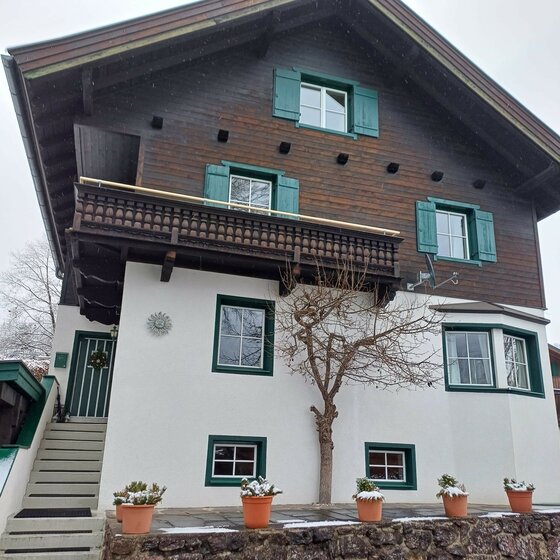 Haus Huber am Sonnenhofweg Alpines Wohnhaus mit weißer Fassade, dunklem Holzobergeschoss, grünem Fensterladen und Balkon.