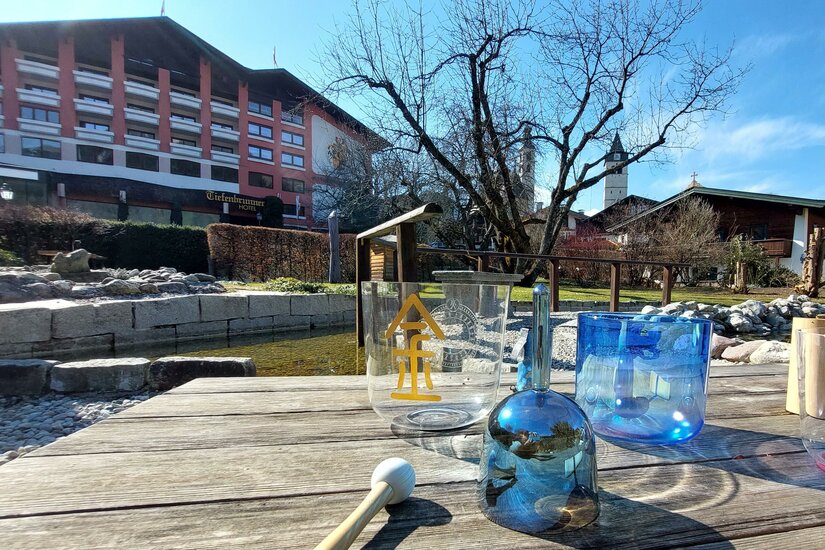 Gläserne Klangschalen auf einem Holztisch im Außenbereich vor einem Hotelgebäude.