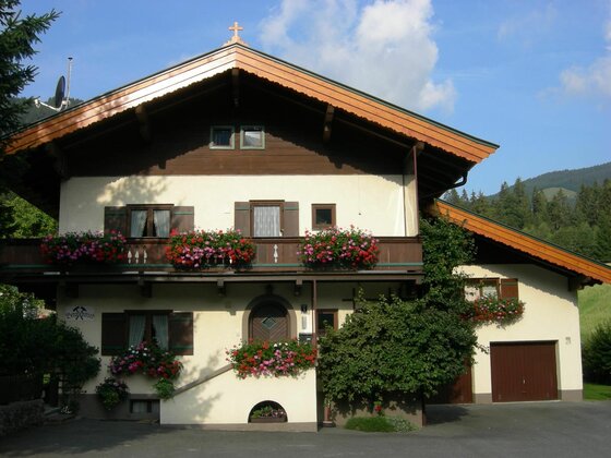 Haus Bergschmied Hausansicht Zweistöckiges Alpenhaus mit Holzbalkon, Blumenschmuck und Satteldach vor bewaldetem Berg.
