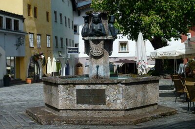 Stadtbrunnen Kitzbühel <p>Ein schöner Brunnen auf einem gemütlichen Platz. Am Brunnen sind Margarethe Maultasch, Ludwig der Strenge und Kaiser Maximilian abgebildet, umgeben von Gebäuden und Terrassen mit Sonnenschirmen.</p>