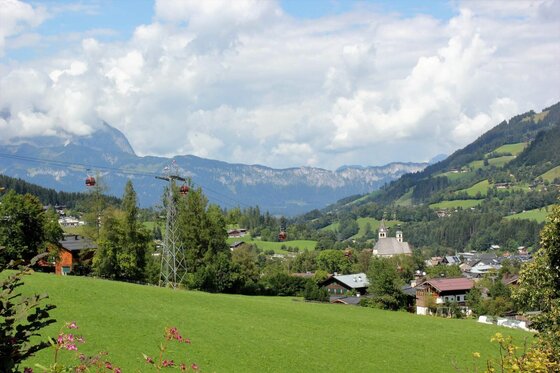 Blick über eine grüne Wiese auf ein Alpendorf mit Seilbahn und Bergen im Hintergrund.