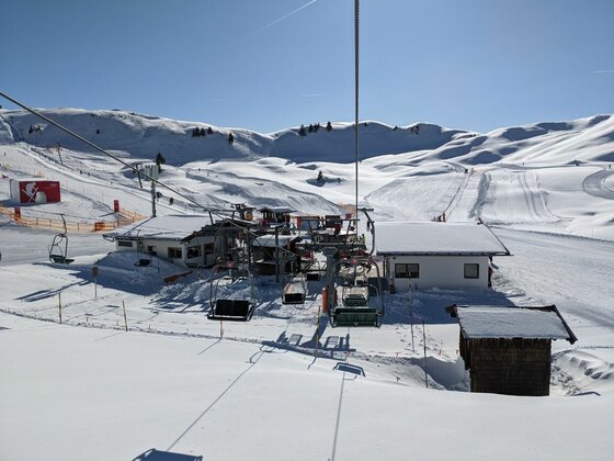 Blick aus einem Sessellift auf eine Talstation in einem verschneiten Skigebiet unter blauem Himmel.