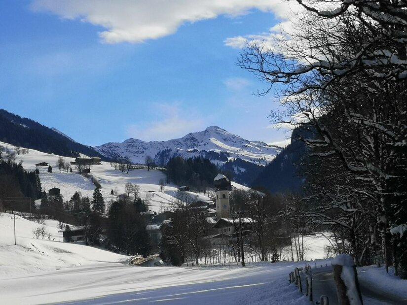 Winterliche Sonnbergrunde Verschneites Bergdorf mit Kirchturm vor schneebedeckten Berggipfeln unter blauem Himmel.