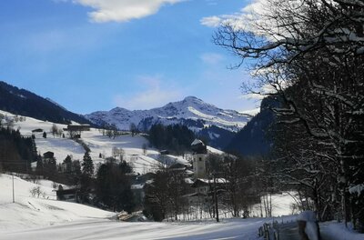 Winterliche Sonnbergrunde Verschneites Bergdorf mit Kirchturm vor schneebedeckten Berggipfeln unter blauem Himmel.