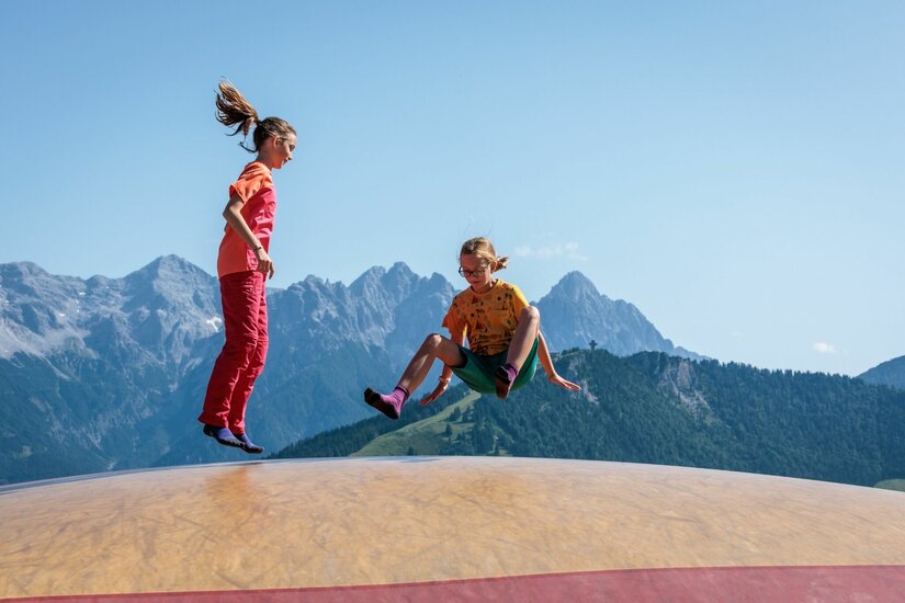 Zwei Kinder springen auf einem großen Hüpfkissen vor einer alpinen Bergkulisse.