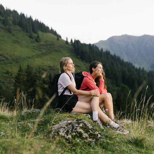 Zwei Frauen sitzen auf einem Felsen in der Natur und genießen die Aussicht auf die grünen Berge und Wälder.