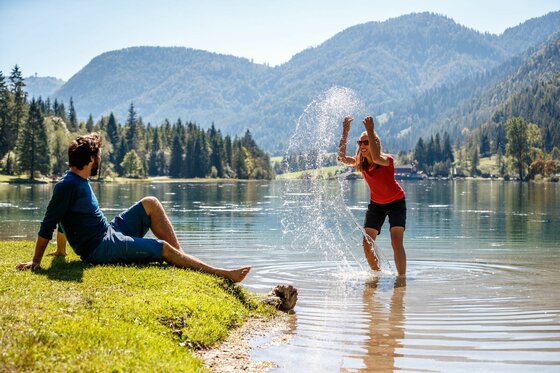 Kat Walk E 6 / E 5 Zwei Personen am Pillersee, eine Frau spritzt im flachen Wasser, ein Mann sitzt am Ufer.