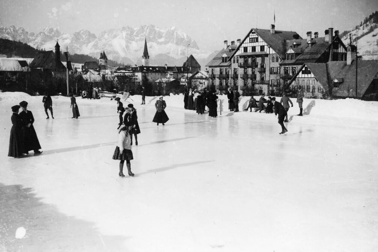 Frauen und Männer beim Eislaufen hinter dem Grand Hotel Kitzbühel, im Hintergrund ist der Wilde Kaiser zu sehen