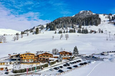 Verschneiter Gasthof mit Parkplatz vor bewaldetem Berghang unter blauem Himmel.