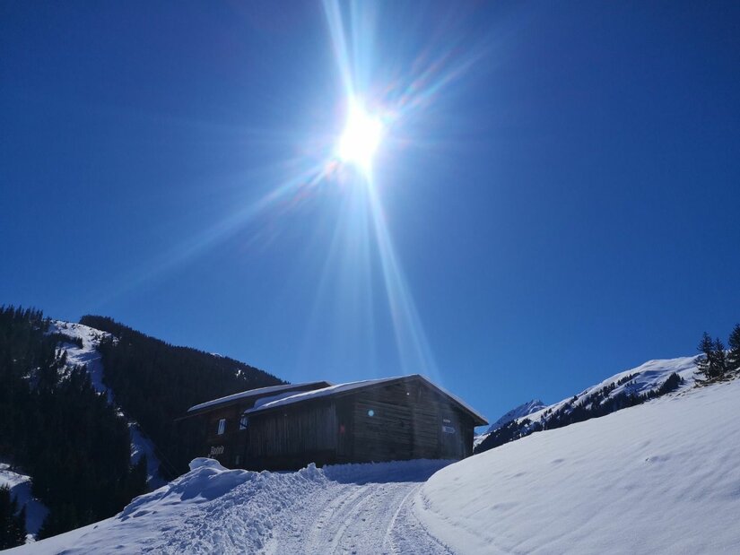 Bruggeralm im Winter Holzhütte in verschneiter Berglandschaft unter strahlend blauem Himmel mit Gegenlicht.