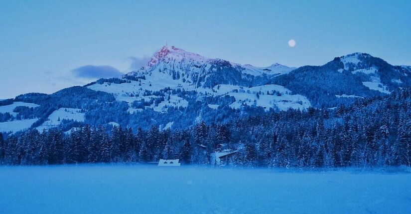 Verschneite Berglandschaft mit Wald und Seeoberfläche in der Dämmerung unter Vollmond.