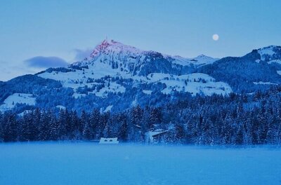 Verschneite Berglandschaft mit Wald und Seeoberfläche in der Dämmerung unter Vollmond.