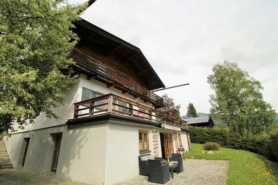 Zweistöckiges alpines Gebäude mit Holzfassade, Balkon und einer Steinterrasse mit Sitzmöbeln.