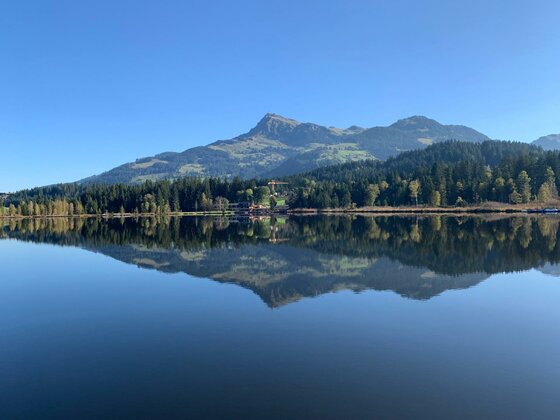 Ein ruhiger Bergsee mit Spiegelung der umliegenden Nadelwälder und Berggipfel unter blauem Himmel.