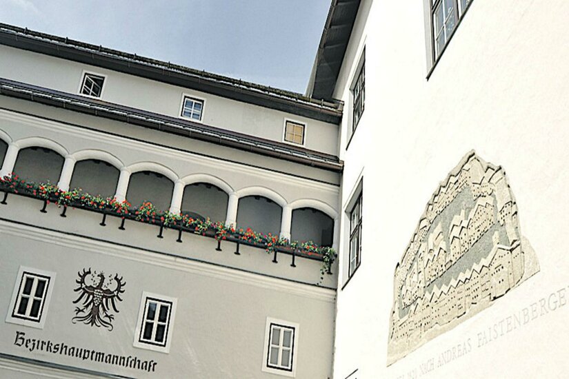 Ein historisches Gebäude mit kunstvollen Fenstern und Blumenkästen. An der Wand ist ein dekoratives Relief zu sehen.
