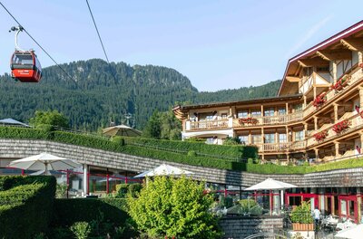 Hotel Kaiserhof und Hahnenkammbahn Rote Seilbahngondel vor bewaldetem Berg und einem Hotelgebäude mit Holzfassade und Blumenschmuck.