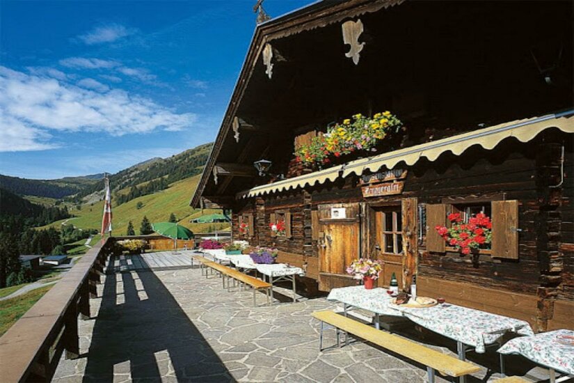 Sonnenterrasse einer rustikalen Holzhütte mit gedeckten Tischen und Blick auf grüne Berghänge.
