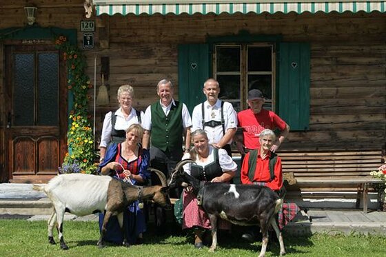 Eine Gruppe von sechs Personen in Tracht steht mit zwei Ziegen vor einem Holzhaus.