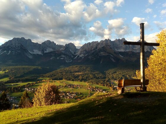 Going_Hollenauer Kreuz_Sommer_2022_4 Eine malerische Berglandschaft mit hohen Gipfeln und buntem Herbstwald. Im Vordergrund steht ein Holzbank mit einem Kreuz, das zur Aussicht einlädt.