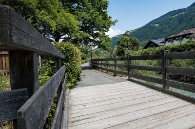 Sonnbergschleife Holzbrücke Holzbrücke führt auf einen asphaltierten Wanderweg entlang eines Zauns vor bewaldeten Bergen.