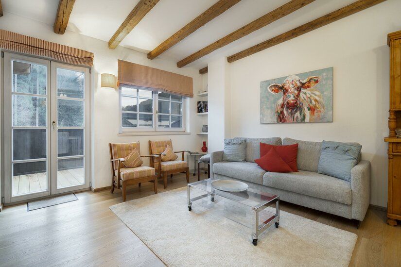 Wohnzimmer mit grauem Sofa, zwei Sesseln, Holzbalkendecke und einem Kuhgemälde an der Wand.