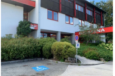 Behindertenparkplatz - Klostergasse 2 Ein modernes Gebäude mit einem Restaurant-Schild. Der Eingangsbereich ist von grünen Pflanzen und einem Behindertenparkplatz umgeben.