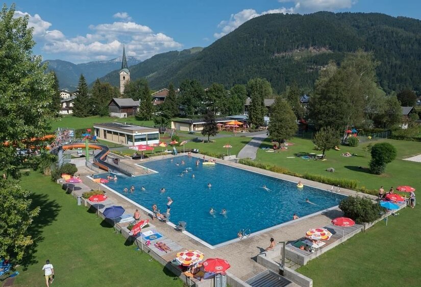 Ein großer Swimmingpool umgeben von grünen Wiesen und Bäumen. Kinder und Erwachsene genießen das Wasser an einem sonnigen Tag.