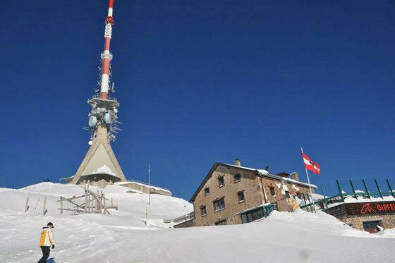 Gipfelhaus im Winter Ein rot-weißer Sendemast und ein steinernes Gipfelhaus in einer verschneiten Gebirgslandschaft unter blauem Himmel.